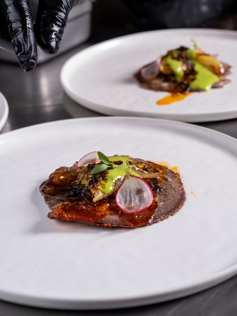 Blue corn tostadas topped with braised meat, avocado crema and radish being plated