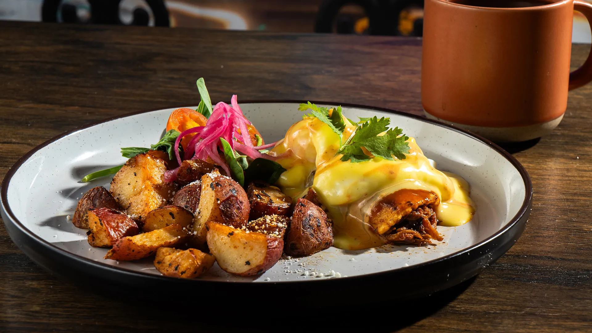 Brunch plate of crispy breakfast potatoes and enchiladas topped with hollandaise and pickled onions, with a cup of coffee